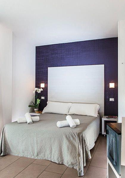 Camera moderna con letto matrimoniale, TV e valigia accanto alla finestra.