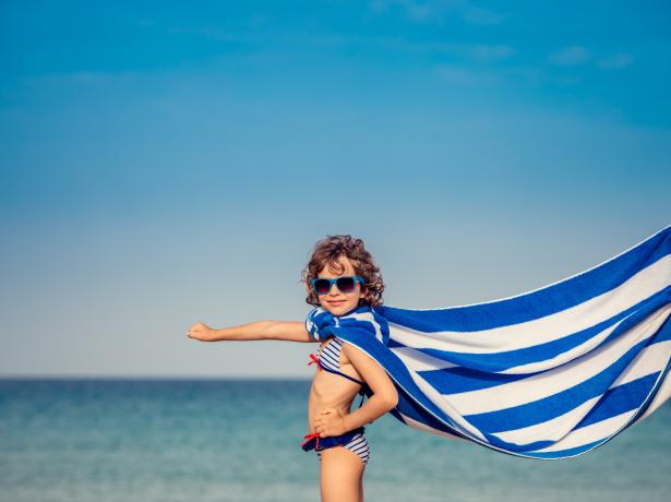 Bambino in costume con occhiali e asciugamano, posa da supereroe sulla spiaggia.