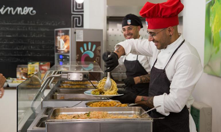 Chefs serving pasta in a cafeteria with smiles.