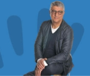 Uomo seduto, occhiali, giacca scura, sfondo blu con forme astratte.