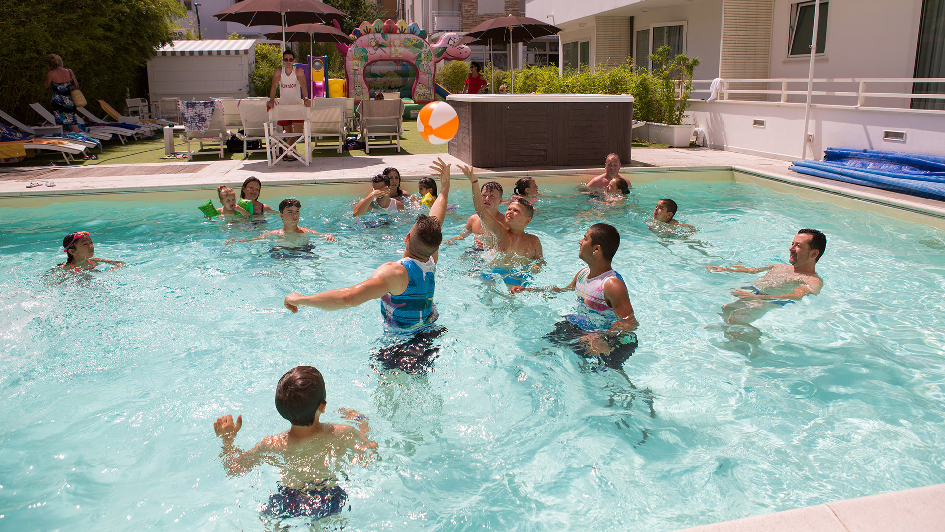 Bambino e uomo in piscina con braccioli, ombrelloni colorati sullo sfondo.