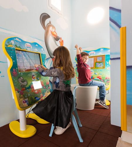 Kinder spielen fröhlich an bunten Bildschirmen, umgeben von Pinguin-Wandbildern.