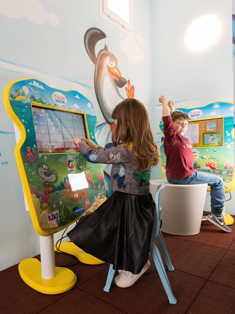 Kinder spielen fröhlich an bunten Bildschirmen, umgeben von Pinguin-Wandbildern.