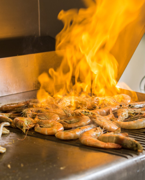 Angolo grill con chef che preparano piatti alla griglia, inclusi gamberi e carne.
